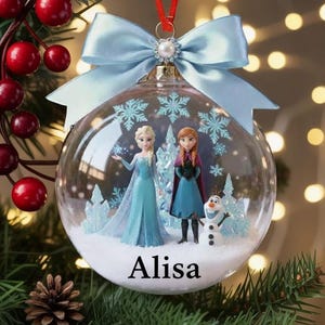 Puede incluir: Una bola de Navidad transparente con un lazo azul claro y una cinta roja. Dentro de la bola hay figuras de Elsa, Anna y Olaf de la película Frozen. El nombre "Alisa" está impreso en la bola.
