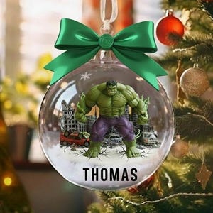 以下が含まれることがあります： ハルクのフィギュアが街のシーンに描かれた透明なガラスのクリスマスオーナメント。オーナメントには緑のリボンと「THOMAS」という名前がプリントされています。オーナメントはクリスマスツリーに飾られています。