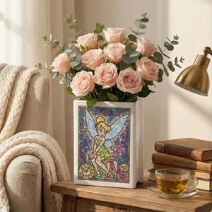 May include: A white rectangular vase with a stained-glass design featuring Tinkerbell, holding a bouquet of pink roses and eucalyptus. The vase sits on a wooden table with books and a teacup.