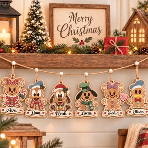 Pode incluir: Um conjunto de seis ornamentos de madeira em forma de personagens de pão de mel pendurados num cordão de juta. Cada ornamento apresenta um design único inspirado em desenhos animados com uma etiqueta de nome: Ava, Liam, Noah, Lucas, Sophia e Oliver.