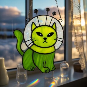 May include: A stained glass cat suncatcher in shades of green, with a white halo and black accents. The cat has a whimsical design with three black antennae. It hangs from a chain, perfect for a window or wall decoration.