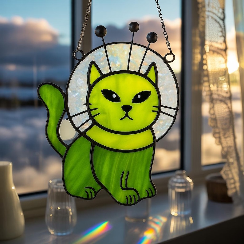 May include: A stained glass cat suncatcher in shades of green, with a white halo and black accents. The cat has a whimsical design with three black antennae. It hangs from a chain, perfect for a window or wall decoration.
