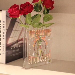 K&ouml;nnte beinhalten: Eine durchsichtige, rechteckige Acrylvase mit drei k&uuml;nstlichen roten Rosen. Die Vorderseite der Vase zeigt einen Grafikdruck mit einem Schwert, Flammen und Stacheldraht sowie den Text "FIRE BREATHING BITCH QUEEN" in einer fetten, stilisierten Schrift.