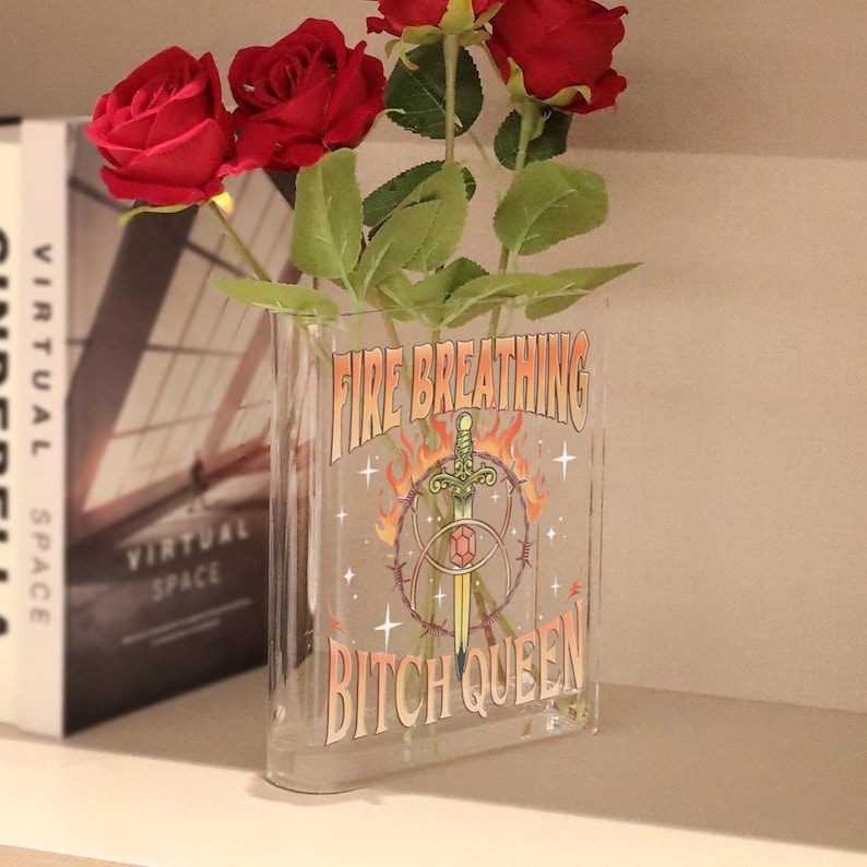 K&ouml;nnte beinhalten: Eine durchsichtige, rechteckige Acrylvase mit drei k&uuml;nstlichen roten Rosen. Die Vorderseite der Vase zeigt einen Grafikdruck mit einem Schwert, Flammen und Stacheldraht sowie den Text "FIRE BREATHING BITCH QUEEN" in einer fetten, stilisierten Schrift.
