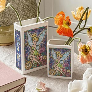 May include: Two white ceramic vases with a stained-glass design featuring Tinkerbell. The vases have a rectangular shape and are decorated with colorful images of the fairy. One vase has the text "Custom Name" on the side. Flowers are placed inside the vases.