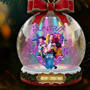 Peut inclure: Une boule de Noël en verre transparent avec un nœud rouge et le texte "HUNTRX". À l'intérieur de la boule se trouvent trois personnages, un chat bleu et l'inscription "MERRY CHRISTMAS" sur une plaque dorée.