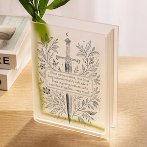 May include: A clear, book-shaped vase featuring a black illustration of a sword, foliage, and text. The text says, "Once upon a time, in a land long since burned to ash, there lived a young princess who loved her kingdom...very much."