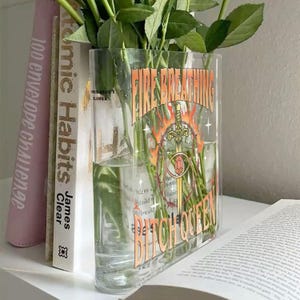K&ouml;nnte beinhalten: Eine rechteckige, durchsichtige Glasvase mit gr&uuml;nen Rosenstielen. Auf der Vorderseite befindet sich ein Grafikdruck eines Schwertes, das von Flammen und Stacheldraht umgeben ist, sowie der Text "FIRE BREATHING BITCH QUEEN" in orangefarbenen Buchstaben.
