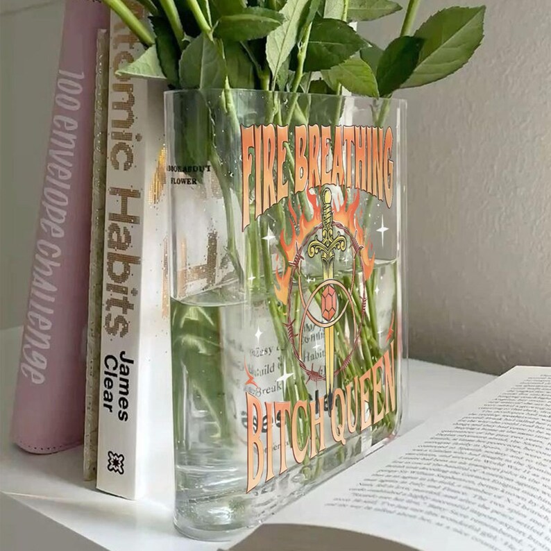 K&ouml;nnte beinhalten: Eine rechteckige, durchsichtige Glasvase mit gr&uuml;nen Rosenstielen. Auf der Vorderseite befindet sich ein Grafikdruck eines Schwertes, das von Flammen und Stacheldraht umgeben ist, sowie der Text "FIRE BREATHING BITCH QUEEN" in orangefarbenen Buchstaben.
