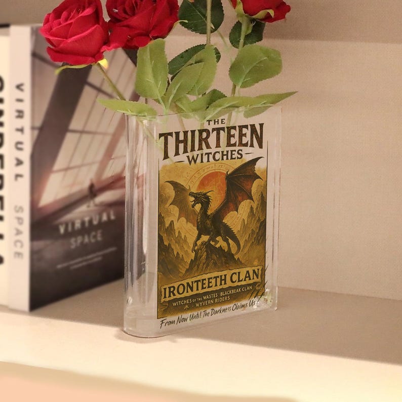 May include: A clear, rectangular glass vase with the text "THE THIRTEEN WITCHES" and "IRONTEETH CLAN" on the front. The vase holds red roses and green leaves. The image has a fantasy theme.