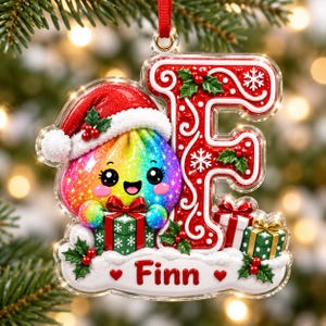 Puede incluir: Un adorno navideño con un personaje de bola de masa de colores del arcoíris con un gorro de Papá Noel rojo, junto a una letra F roja grande decorada con remolinos blancos y copos de nieve. El nombre Finn aparece impreso abajo con regalos.