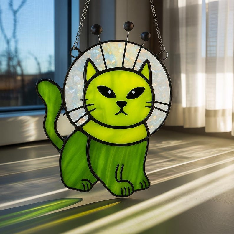 May include: A stained glass cat suncatcher in shades of green, with a white halo and black accents. The cat has large, black eyes and a whimsical design. It hangs from a chain and is displayed in front of a window.
