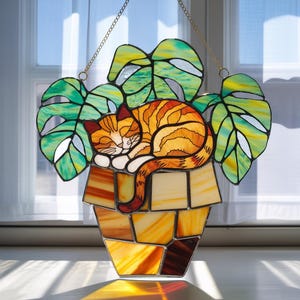 May include: Stained glass window art featuring a sleeping orange tabby cat nestled in a pot with green monstera leaves. The cat is curled up, and the pot is made of various shades of orange and yellow glass. A gold chain is attached for hanging.