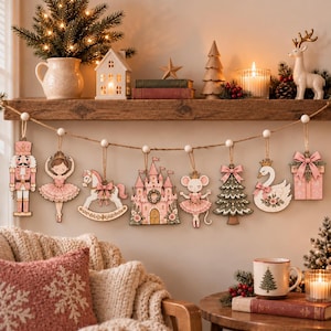 Op de afbeelding: Een decoratieve slinger met acht houten ornamenten in zachte roze en gedempte tinten, waaronder een notenkraker, ballerina, hobbelpaard, kasteel, muis, kerstboom, zwaan en cadeau, allemaal opgehangen aan een natuurlijk touw van jute.
