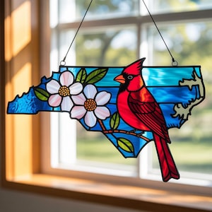 May include: Stained glass art piece in the shape of North Carolina, featuring a red cardinal bird perched on a branch with white and pink dogwood flowers. The background is blue with a water-like pattern.