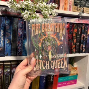 K&ouml;nnte beinhalten: Eine rechteckige, durchsichtige Acrylvase mit wei&szlig;en Schleierkrautbl&uuml;ten. Die Vorderseite zeigt einen Grafikdruck mit dem Text "FIRE BREATHING BITCH QUEEN" in beiger Schrift, der eine Schwertillustration mit einem roten Edelstein umrahmt.