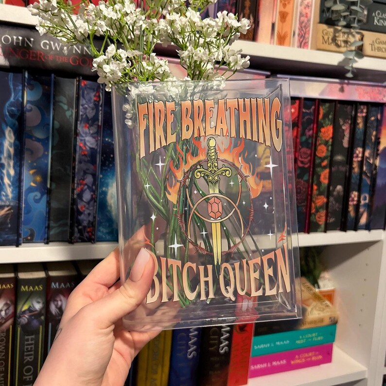 K&ouml;nnte beinhalten: Eine rechteckige, durchsichtige Acrylvase mit wei&szlig;en Schleierkrautbl&uuml;ten. Die Vorderseite zeigt einen Grafikdruck mit dem Text "FIRE BREATHING BITCH QUEEN" in beiger Schrift, der eine Schwertillustration mit einem roten Edelstein umrahmt.