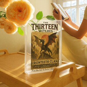 May include: A clear acrylic book display case holding a book titled "The Thirteen Witches." The book cover features a black dragon illustration with the words "Ironteeth Clan." The case sits on a wooden tray with a bouquet of orange roses.