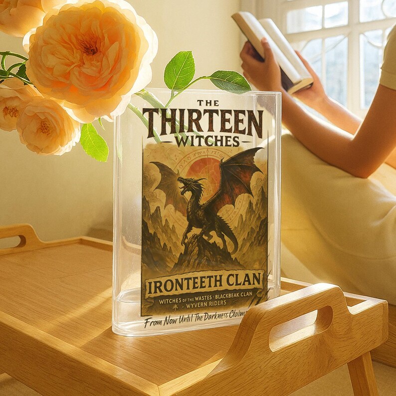 May include: A clear acrylic book display case holding a book titled "The Thirteen Witches." The book cover features a black dragon illustration with the words "Ironteeth Clan." The case sits on a wooden tray with a bouquet of orange roses.