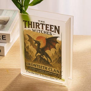 May include: A clear, book-shaped vase with the title "The Thirteen Witches" and a dragon illustration. The vase also features the text "Ironteeth Clan" and "From Now Until The Darkness Claims Us". Green plant stems are visible inside the vase.