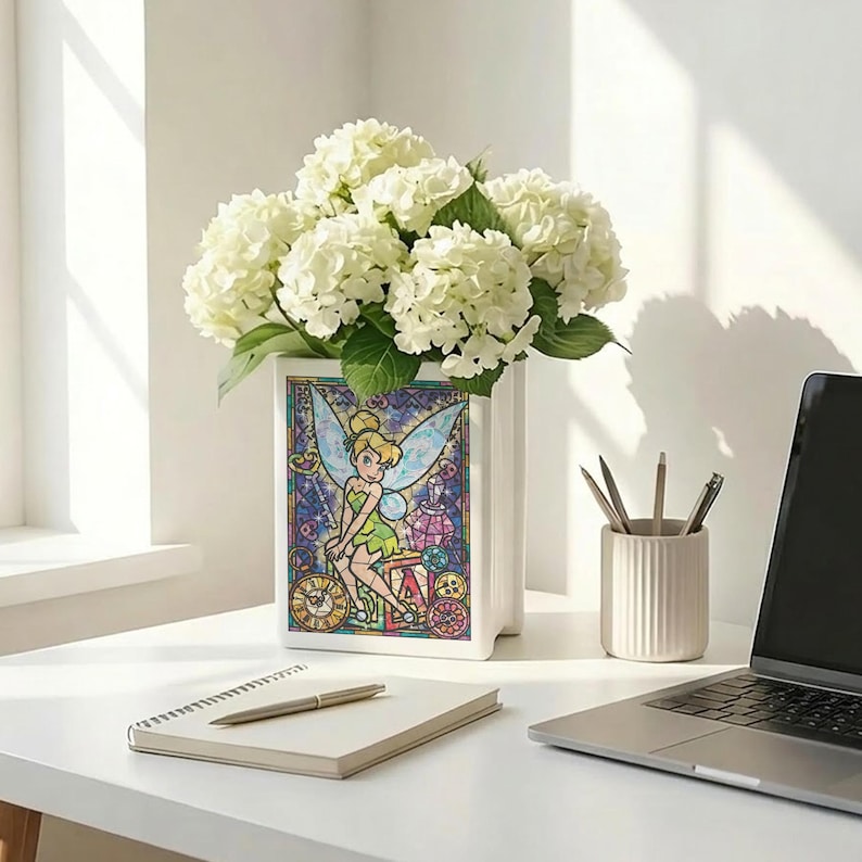 May include: A white ceramic vase with a stained-glass design featuring Tinkerbell, holding a bouquet of white hydrangeas. The vase is on a white desk with a notebook, pen, and laptop.