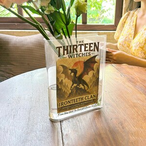 May include: A clear, rectangular vase holding flowers and a book titled "The Thirteen Witches." The book cover features a dragon illustration with the words "Ironteeth Clan" and "From Now Until The Darkness Claims Us."