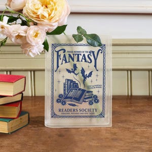 May include: A clear, book-shaped vase with blue text that reads "PROUD MEMBER OF THE FANTASY READERS SOCIETY". The design includes bats, books, and the phrase "LOST IN FANTASY FOUND IN BOOKS". A bouquet of flowers sits behind the vase.