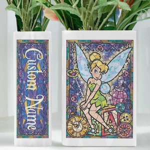May include: Two white ceramic vases with a stained-glass design. One vase features the text "Custom Name" in gold lettering. The other vase has a colorful image of Tinkerbell from Peter Pan. The vases are filled with greenery.