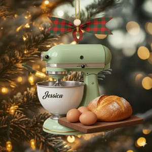 May include: A mint green stand mixer ornament with a white bowl that has the name "Jessica" printed on it. The ornament is decorated with a red and green plaid bow and a gold string. A loaf of bread and three eggs sit on a wooden cutting board.
