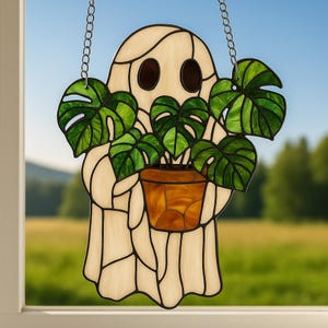 May include: A stained glass ghost holding a potted plant with large green leaves. The ghost is off-white with black outlines and brown eyes. The plant pot is brown and orange. A silver chain is attached to the top for hanging.