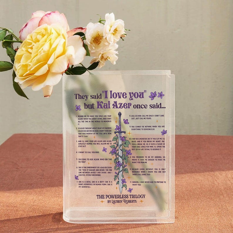 May include: A clear acrylic bookend featuring a quote from the book "The Powerless Trilogy" by Lauren Roberts. The quote reads "They said "I love you" but Kai Azer once said..." and is followed by a list of 12 statements. The bookend features a purple and green floral design with a sword.