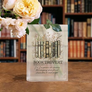 May include: A clear, rectangular vase with the word "BOOKTROVERT" and its definition printed on the front. The design features a collection of vintage books and floral accents. A bouquet of yellow and white roses sits in front of the vase.
