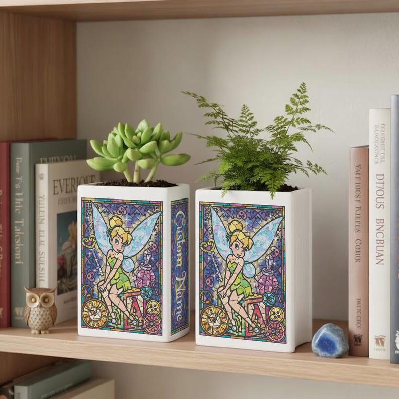 May include: Two white rectangular planters with a stained-glass design featuring Tinkerbell. The planters hold green plants and have the text "Custom Name" on the side. The planters are on a wooden shelf with books and decorative items.