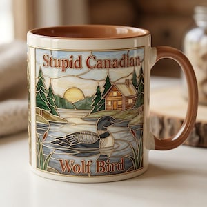 May include: A ceramic mug with a brown interior and handle, featuring a stained-glass style illustration of a lake, a cabin, pine trees, and a loon. The text "Stupid Canadian" is printed at the top and "Wolf Bird" is printed at the bottom.