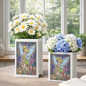 May include: Two white ceramic vases with a stained-glass design featuring Tinkerbell. One vase holds white daisies, the other holds blue and white hydrangeas. The vases are on a wooden table.