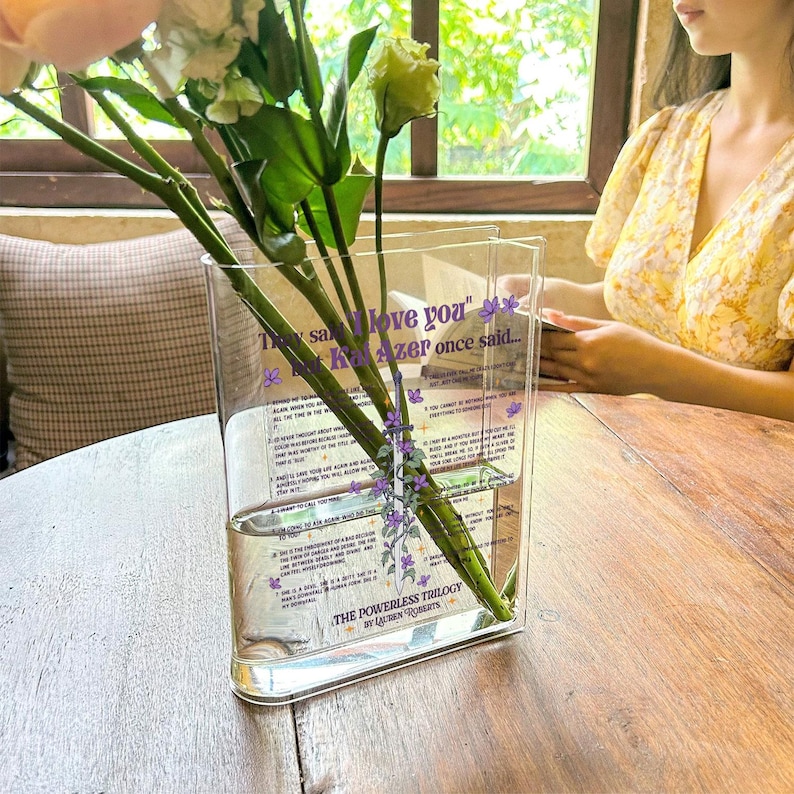 Op de afbeelding: Een heldere glazen vaas met een paars en wit bloemenontwerp. De vaas is gevuld met water en bloemen. De vaas heeft tekst die luidt "Ze zeggen dat ik van je hou" maar Kaz Azer zei ooit..." en "The Powerless Trilogy by Leigh Bardugo".