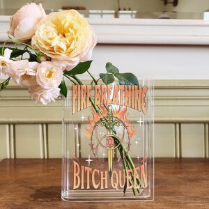 K&ouml;nnte beinhalten: Eine rechteckige, transparente Acrylvase mit einem Grafikdruck aus Schwert, Flammen und Stacheldraht sowie dem Text FIRE BREATHING BITCH QUEEN. Die Vase enth&auml;lt mehrere blassrosa und cremefarbene Rosen mit gr&uuml;nen Stielen und Bl&auml;ttern.