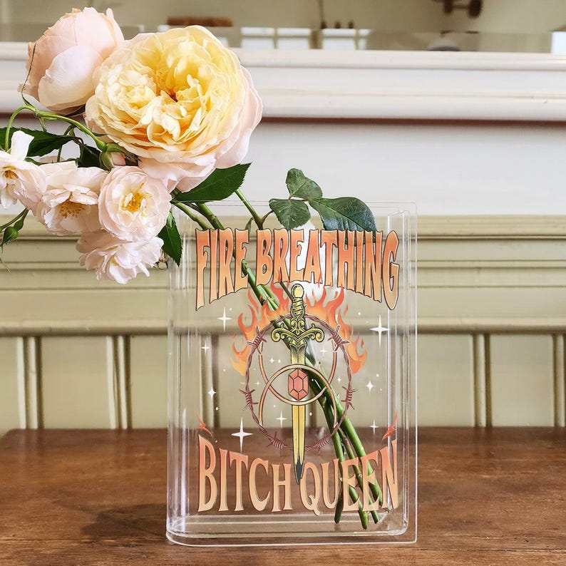 K&ouml;nnte beinhalten: Eine rechteckige, transparente Acrylvase mit einem Grafikdruck aus Schwert, Flammen und Stacheldraht sowie dem Text FIRE BREATHING BITCH QUEEN. Die Vase enth&auml;lt mehrere blassrosa und cremefarbene Rosen mit gr&uuml;nen Stielen und Bl&auml;ttern.
