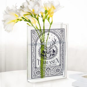 May include: A clear rectangular vase with a decorative black and white graphic of a stag, mountains, and the text "TERRASEN HOME OF THE STAG." The vase holds white and yellow flowers.