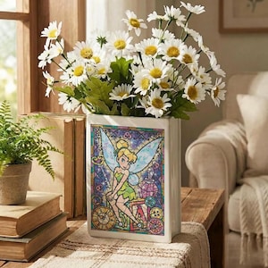 May include: A white rectangular vase with a stained-glass design featuring Tinkerbell. The vase holds a bouquet of white daisies with yellow centers and green leaves. The vase is on a wooden table with books and a small potted fern.