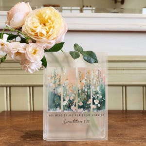 May include: A clear glass vase with a floral design and the text "His mercies are new every morning. Lamentations 3:23". The vase is filled with pale pink and yellow roses.