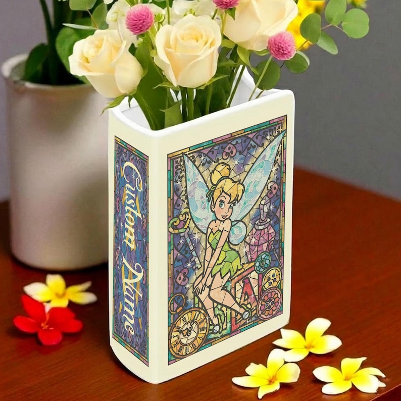 May include: A cream-colored ceramic vase shaped like a book, featuring a stained-glass style illustration of Tinkerbell. The vase has the words "Custom Name" on the side and is filled with white and pink flowers. Yellow flowers are scattered on the table.