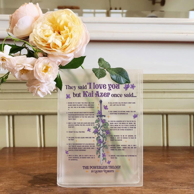 Op de afbeelding: Een witte, gematteerde glazen plaquette met een paars en groen bloemenmotief en een citaat uit het boek "The Powerless Trilogy" van Lauren Roberts. Het citaat luidt "Ze zeiden "Ik hou van je" maar Kai Azer zei ooit..." en somt 12 redenen op.