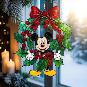 Puede incluir: Un adorno navideño de Mickey Mouse. El adorno presenta a Mickey Mouse en el centro, rodeado por una corona de hojas verdes de acebo y bayas rojas. Un lazo rojo se encuentra en la parte superior. El adorno tiene una apariencia festiva y alegre.