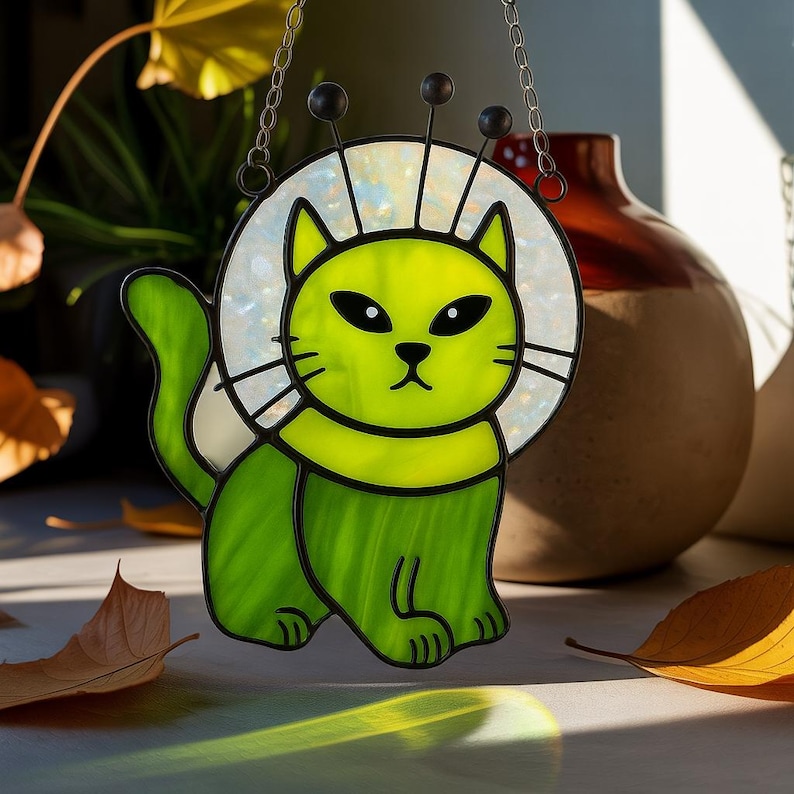 May include: A stained glass cat suncatcher in shades of green, with a white and iridescent halo. The cat has black eyes and whiskers, and is suspended by a silver chain. The cat has three black antennae.