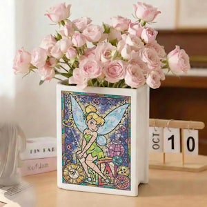 May include: A white ceramic vase shaped like a book, featuring a stained-glass design of Tinkerbell. The vase holds a bouquet of pink roses. The design includes clocks and gears. A wooden calendar shows October 10.