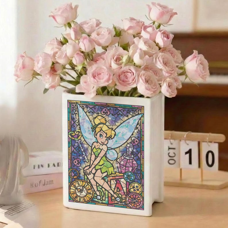 May include: A white ceramic vase shaped like a book, featuring a stained-glass design of Tinkerbell. The vase holds a bouquet of pink roses. The design includes clocks and gears. A wooden calendar shows October 10.