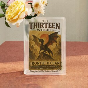 May include: A clear acrylic book display case featuring the title "THE THIRTEEN WITCHES" and a dragon illustration. The case also displays "IRONTEETH CLAN" and the phrase "From Now Until The Darkness Claims Us". Yellow roses and white flowers are in the background.