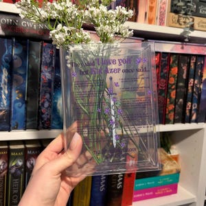 Op de afbeelding: Een doorzichtige plastic vaas met een citaat uit de Throne of Glass-serie van Sarah J. Maas. Het citaat luidt: "Ze zeiden 'Ik hou van je', maar Kaz Brekker zei ooit..." De vaas is gevuld met witte bloemen en heeft een paarse en witte illustratie van een zwaard en wijnstokken.