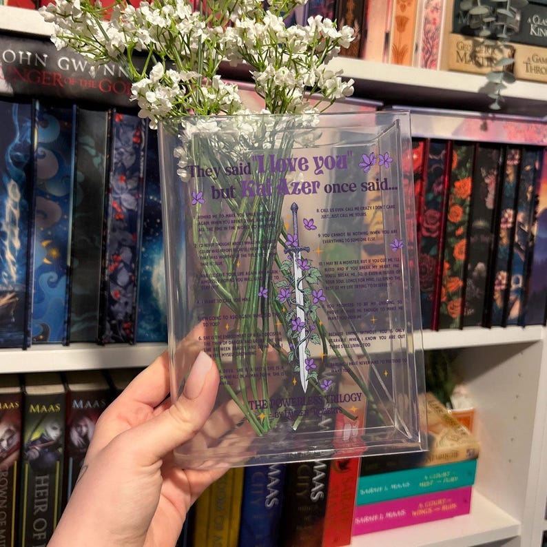 Op de afbeelding: Een doorzichtige plastic vaas met een citaat uit de Throne of Glass-serie van Sarah J. Maas. Het citaat luidt: "Ze zeiden 'Ik hou van je', maar Kaz Brekker zei ooit..." De vaas is gevuld met witte bloemen en heeft een paarse en witte illustratie van een zwaard en wijnstokken.